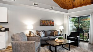 Un salon moderne avec un canapé gris, un fauteuil à motifs et une table basse. Un tableau orne le mur et une grande fenêtre offre une vue sur l’extérieur.