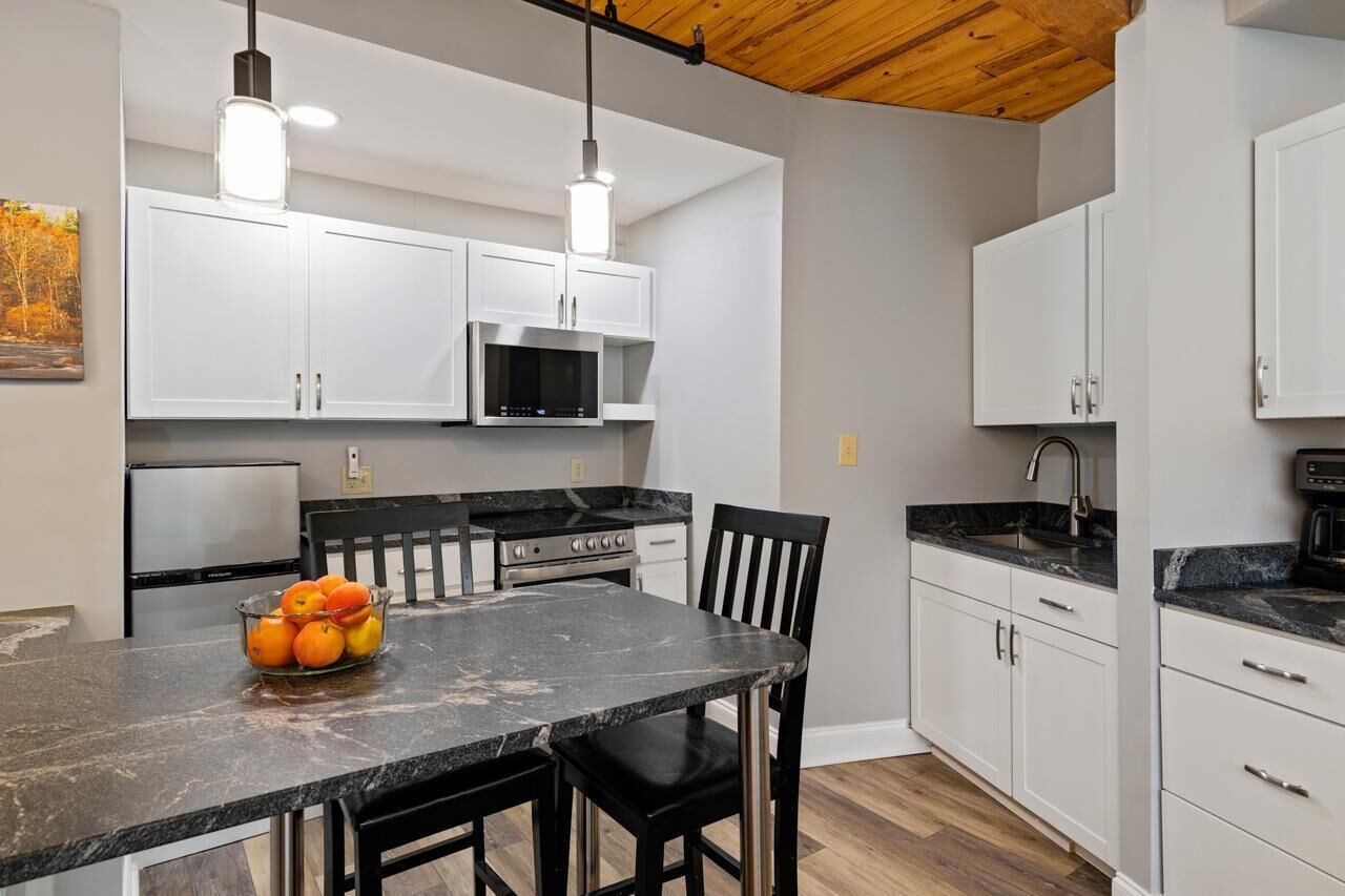 A modern kitchen with white cabinets, a black countertop, and a dining area with a black chair and a fruit bowl.