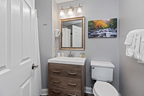 A bathroom with a white sink, wooden cabinet, and a toilet. There is a mirror with a picture of a river and autumn leaves on the wall.