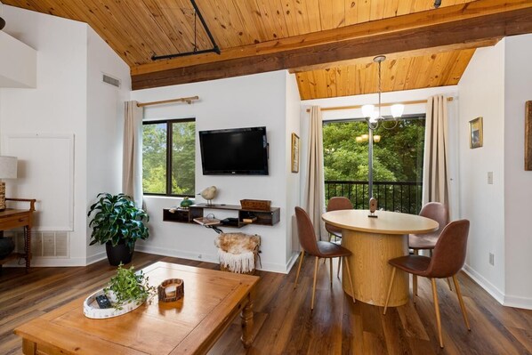 Un salón moderno con techo de madera, una mesa redonda de comedor con cuatro sillas, un televisor montado en la pared y ventanas grandes con vistas a la vegetación.