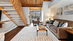 Ein modernes Wohnzimmer mit einer Holztreppe, einem Sofa, einem Couchtisch und Blick durch ein großes Fenster.