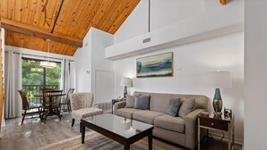 Un salon spacieux avec un plafond en bois, un canapé beige, une table basse en verre et un coin repas comprenant une table et des chaises.