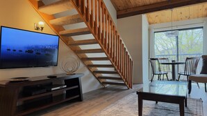 Un salon moderne avec un escalier en bois, un grand téléviseur à écran plat, une table à manger en verre et une vue sur l’extérieur.