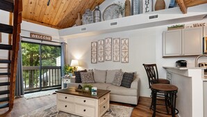 A modern living room with a sofa, coffee table, and decorative pillows. A kitchen area with cabinets and a sink is visible to the right.