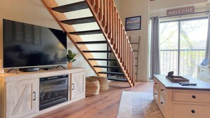 A modern living room with a large flat-screen TV, a wooden staircase, and a 'Welcome' sign near a large window.