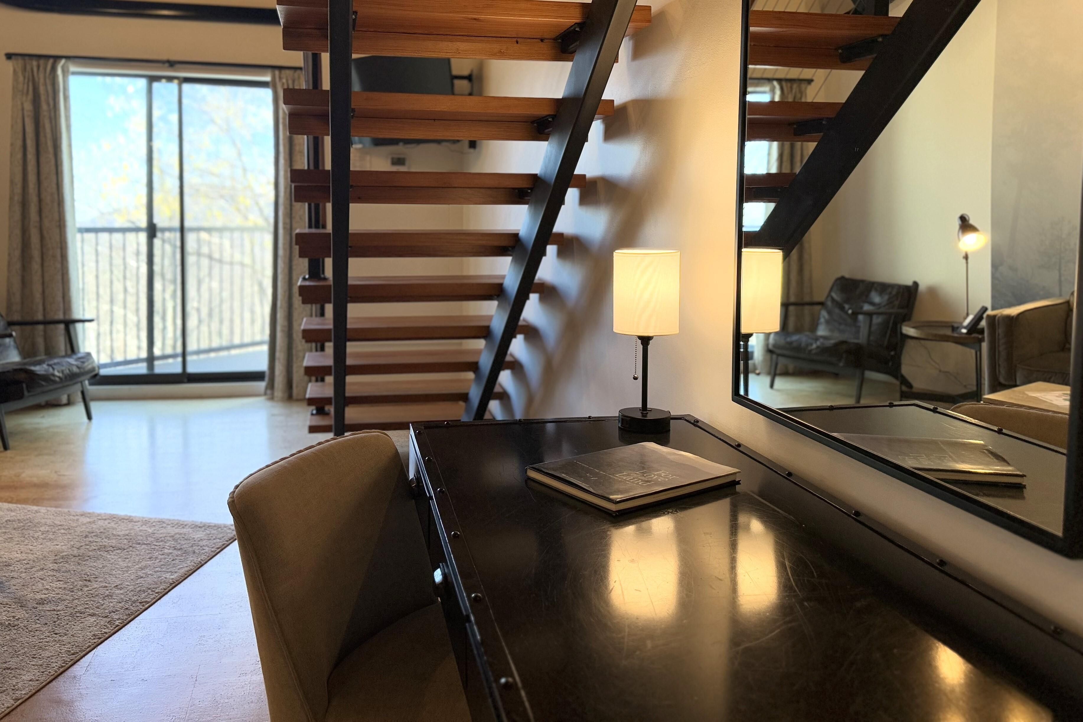 Une pièce moderne avec une table en bois foncé, un fauteuil en cuir, une lampe et un grand miroir qui reflète un escalier et un fauteuil.