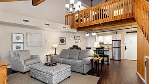Un vaste espace de vie comprenant un canapé gris, un pouf assorti et une table basse en bois. Un escalier avec une rampe en bois mène à l’étage supérieur. On aperçoit la cuisine à l’arrière-plan, équipée d’appareils électroménagers modernes.