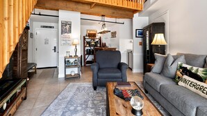 Un amplio salón con un sofá gris, una mesa de centro de madera y un sillón negro. Una escalera de madera conduce a un nivel superior.
