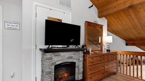 Una sala de estar acogedora con chimenea de piedra, televisor, cómoda de madera y una escalera que sube a un nivel superior.