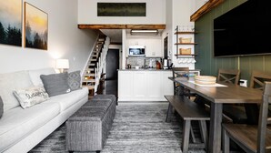 Una sala moderna con un sofá, una mesa de centro y una mesa de comedor con sillas. Hay una zona de cocina con un microondas y armarios. Se ve una escalera al fondo.