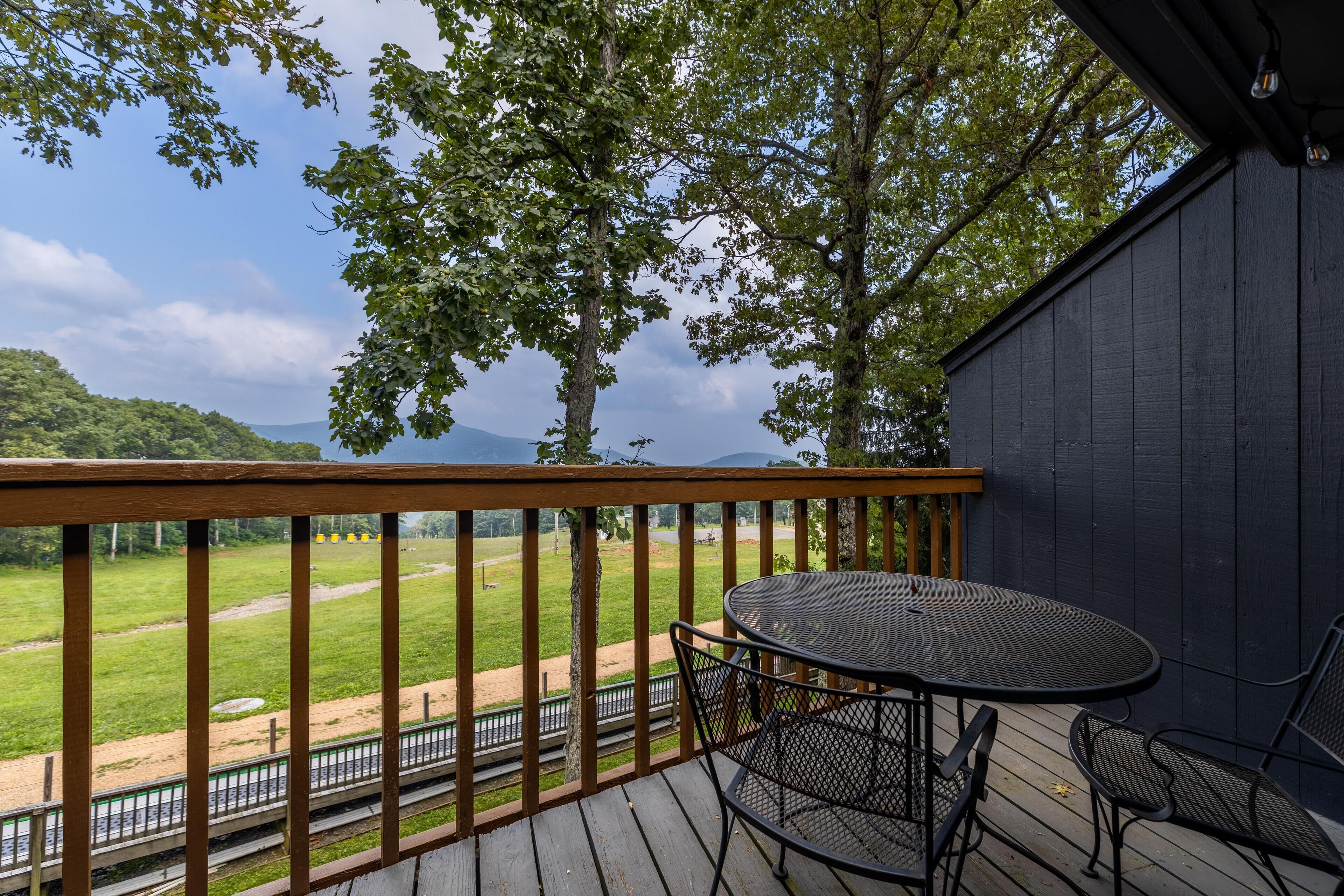 A wooden deck with a metal table and chairs, overlooking a green field and distant mountains.