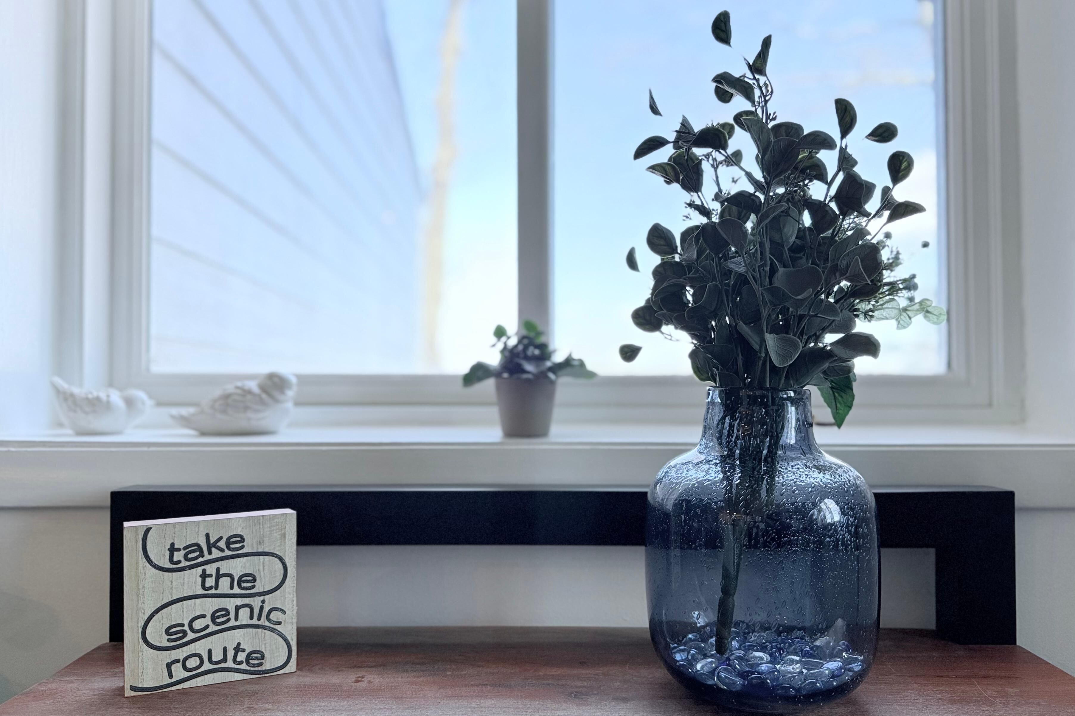 A clear view of a window with a white blind, a small potted plant on the windowsill, and a wooden table with a clear glass vase containing green foliage and a sign that reads "take the scenic route."