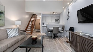 Una sala moderna con sofá, mesa de centro y televisor. Se ve una escalera que lleva a una zona de cocina con mesa y sillas.