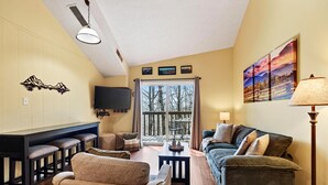 Un salón con sofá, sillones, una mesa de centro, televisor y un balcón con vista a árboles.