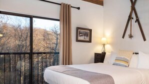 A bedroom with a large bed, a nightstand, a lamp, a picture on the wall, and a view of trees through a balcony. - Mountain Inn Studio 53 with Wintergreen Advantage (Wintergreen)