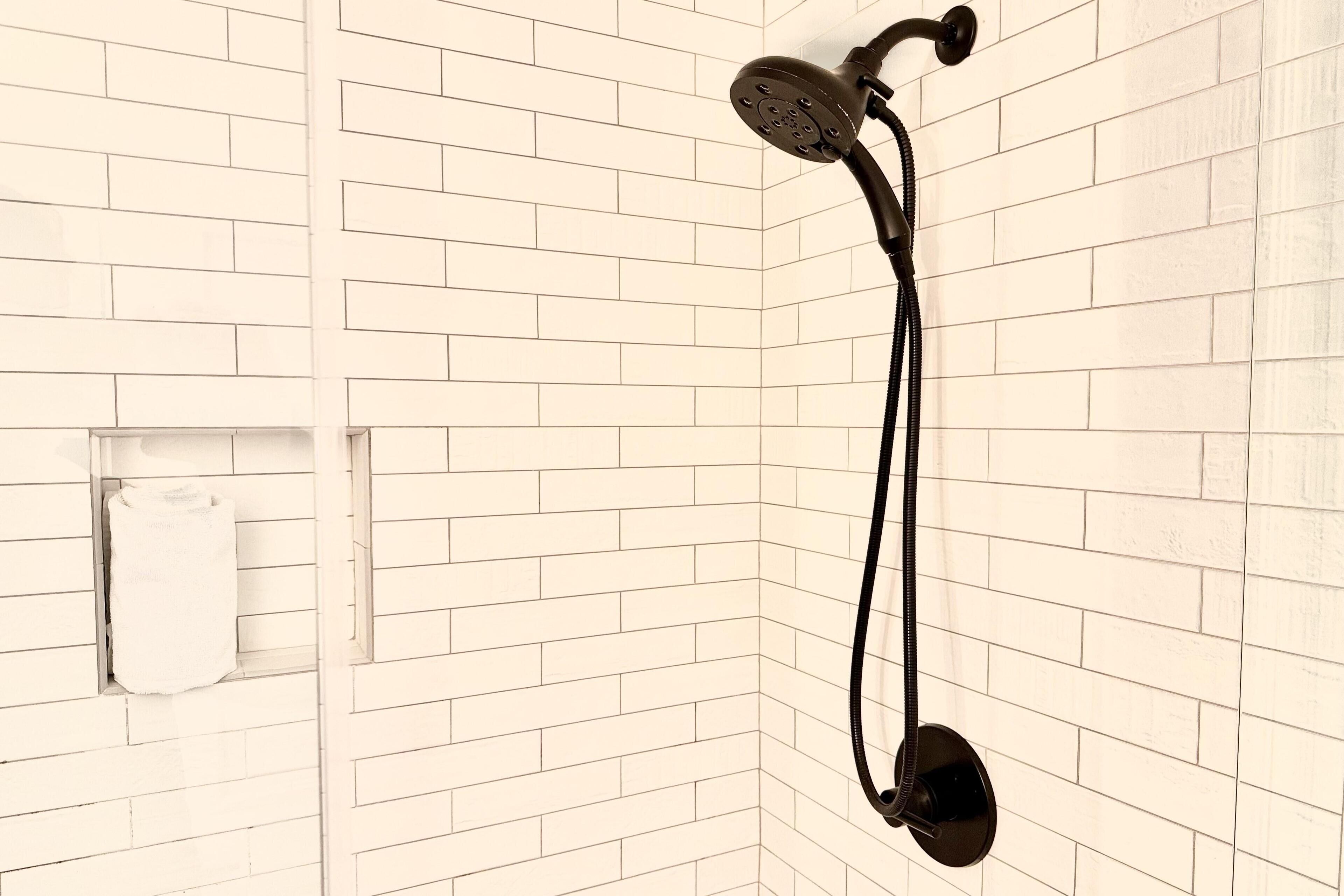 A corner shower with a handheld showerhead and a handheld scrubber, mounted on a white tiled wall.