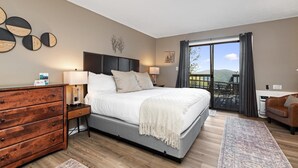 A bedroom with a bed, wooden dresser, bedside tables, a balcony with a view, and a sofa.