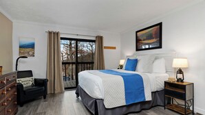 Ein Schlafzimmer mit einem großen Bett, einem Nachttisch mit Lampe, einem Schminktisch, einem Sessel und einem Fenster mit Vorhängen.