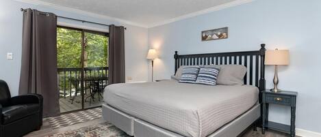 A bedroom with a large bed, a nightstand, a chair, a window with curtains, and a view of greenery outside.