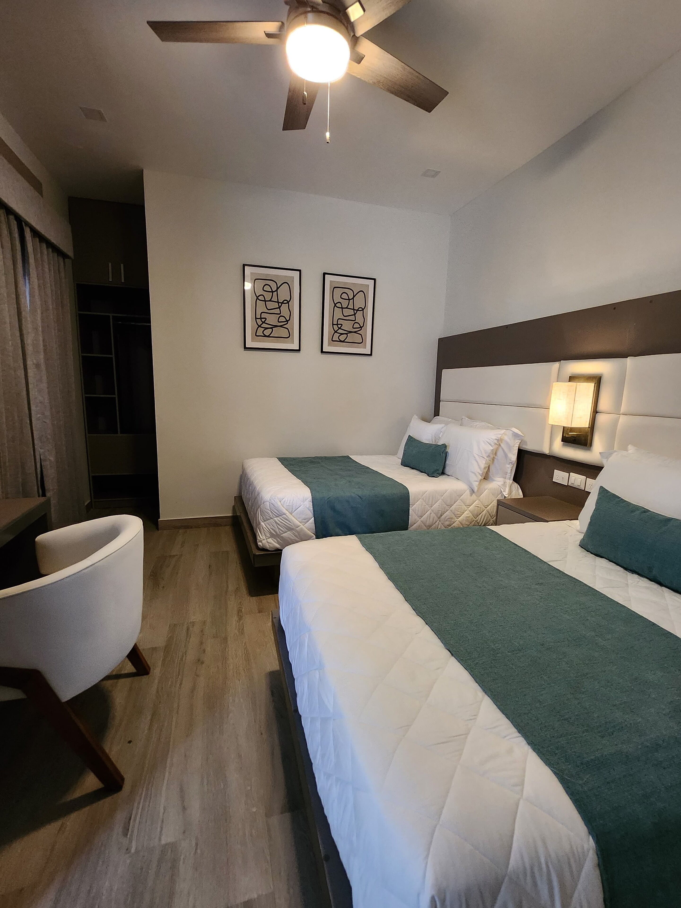 Quarto de hotel com duas camas, um ventilador de teto, uma escrivaninha e uma cadeira.