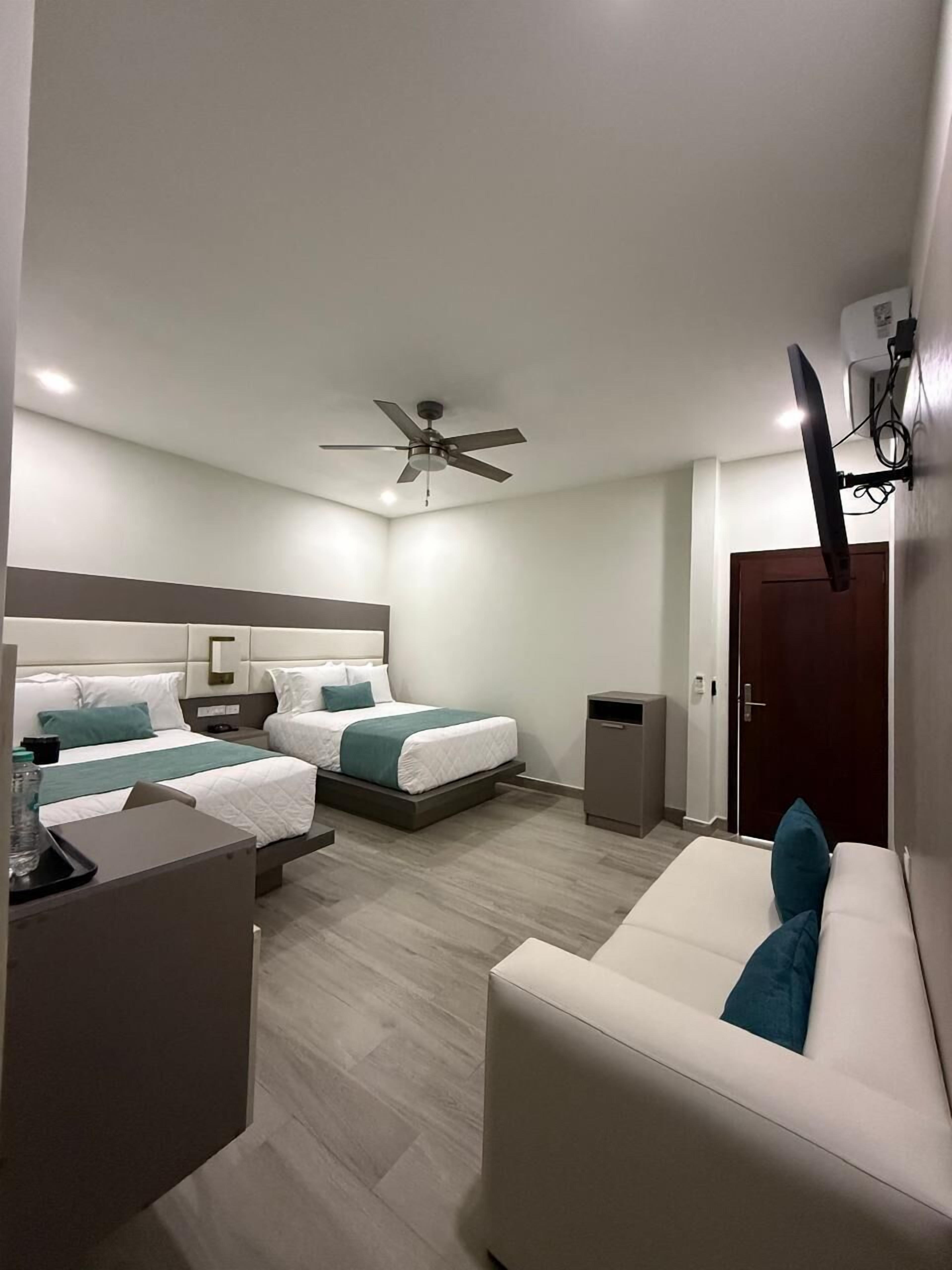 Una habitación de hotel moderna con dos camas, un ventilador de techo, un televisor montado en la pared y una mesita con una silla.