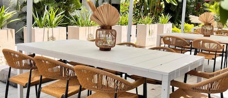 Un espacio exterior para comer con mesas blancas y sillas de mimbre, rodeado de abundante follaje y plantas en macetas.