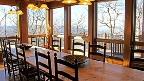 Une table à manger en bois, des chaises, de grandes fenêtres et des lampes suspendues.