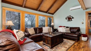 Un salón amplio con sofás de cuero, una mesa de centro de madera y ventanas grandes con vistas a árboles.