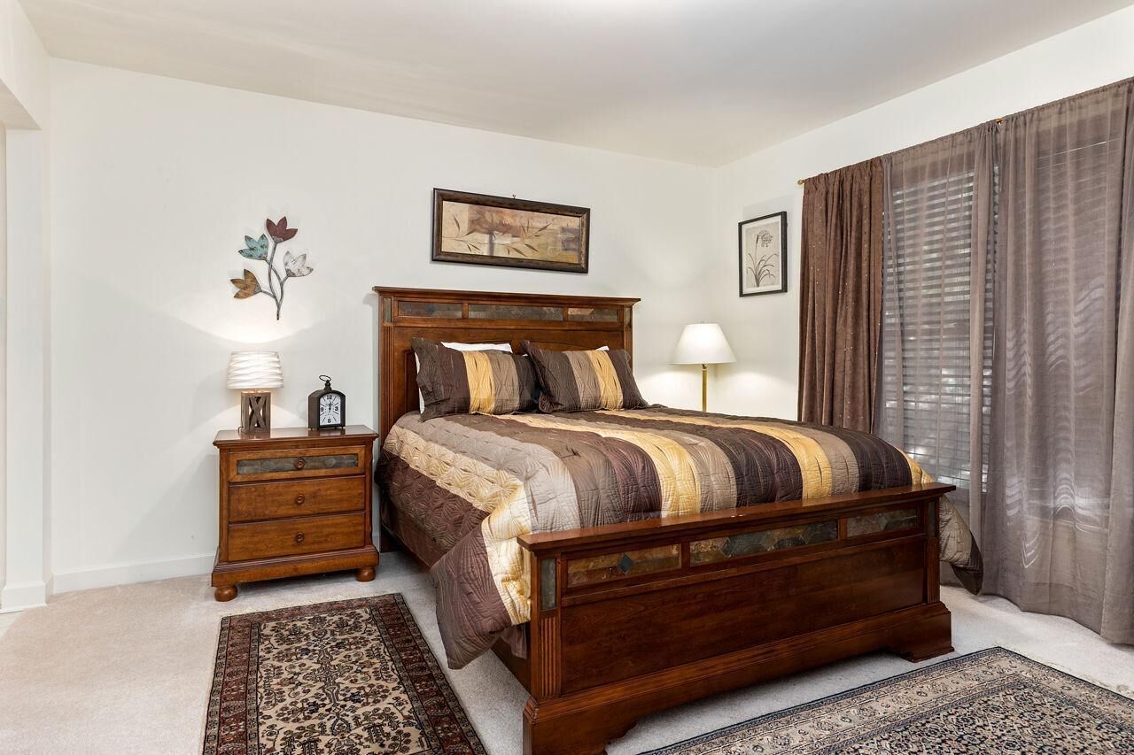 A bedroom with a wooden bed, two bedside tables, a nightstand, a large rug, and a window with curtains.