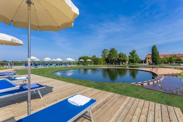 A pool area with lounge chairs and umbrellas, surrounded by greenery and a clear sky.