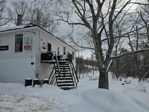 A white building with a staircase leading to a door, surrounded by snow-covered ground and trees. - Charming Historic Loft Apartment in Waldoboro. Once an Army Post & Dance Hall. (Waldoboro)