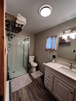 A bathroom with a glass-enclosed shower, a toilet, a sink with a mirror, and towels on a rack.