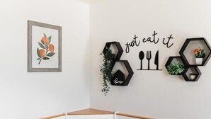 Un comedor con una mesa de madera y sillas blancas, una obra de arte enmarcada con frutas, y una decoración de pared con utensilios y la frase 