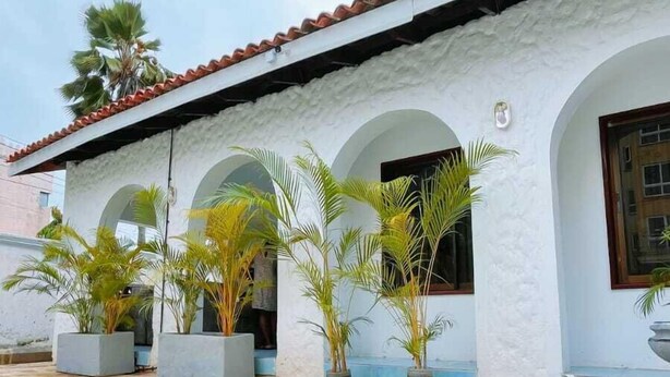 Un bâtiment à l’extérieur blanc, avec un toit recouvert de tuiles rouges et de nombreuses arcades. On y trouve des palmiers en pot et une cour dallée de pierres.