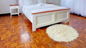 Une chambre avec un parquet en bois, un lit blanc doté d’une tête de lit en bois, une table de chevet blanche et un ventilateur de plafond.