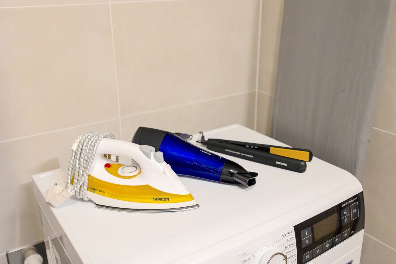 Un fer à repasser blanc et un sèche-cheveux bleu sont posés sur un appareil blanc, qui semble être un fer à vapeur, dans une salle de bain dont les murs sont recouverts de carreaux.
