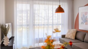 Un salon avec un canapé, une suspension décorative, une grande fenêtre avec des rideaux légers et un tableau encadré accroché au mur.
