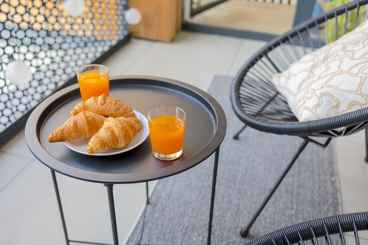 A breakfast tray with croissants and orange juice on a small table, set on an outdoor patio with a wicker chair and a patterned cushion.