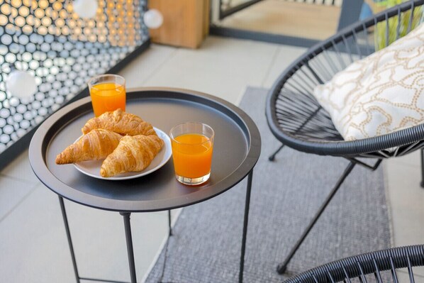 A breakfast tray with croissants and orange juice on a small table, set on an outdoor patio with a wicker chair and a patterned cushion.