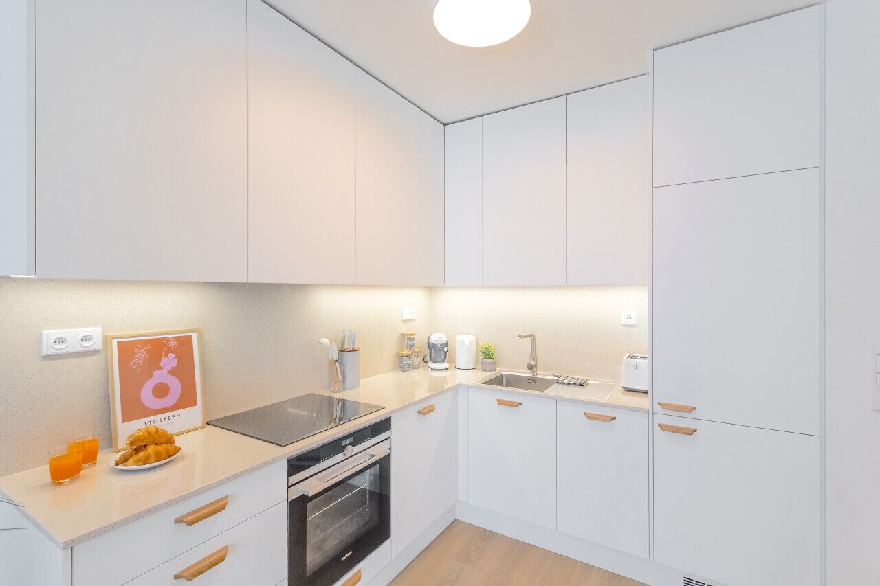 Eine moderne Küche mit weißen Schränken, einem eingebauten Backofen und einer Spüle. An der Wand hängt ein Poster und auf der Arbeitsfläche liegt eine Platte mit Croissants.