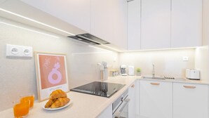 Une cuisine moderne avec des meubles blancs, une plaque de cuisson à induction noire et un évier. On peut voir deux verres de jus d’orange et une assiette de croissants sur le comptoir.