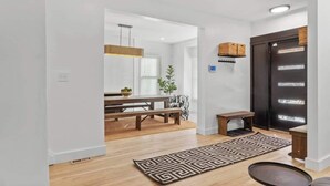 A modern interior with a wooden floor, a patterned rug, a bench, a dining table with chairs, and a dark door with a basket. - Spacious Detroit Retreat • Sleeps 16 • Parking (Detroit)
