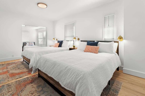 A bedroom with two beds, white bedding, patterned rugs, and wall-mounted lamps.