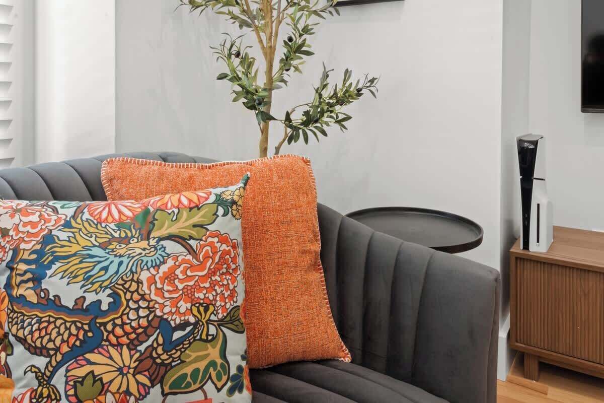 Un salon moderne avec un canapé gris, un coussin décoratif coloré à motifs brodés et une table d’appoint en bois sur laquelle se trouve un objet noir.