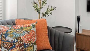 Un salon moderne avec un canapé gris, un coussin décoratif coloré à motifs brodés et une table d’appoint en bois sur laquelle se trouve un objet noir.