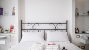 A neatly made bed with white linens and towels, a metal headboard, and shelves with books and toiletries.