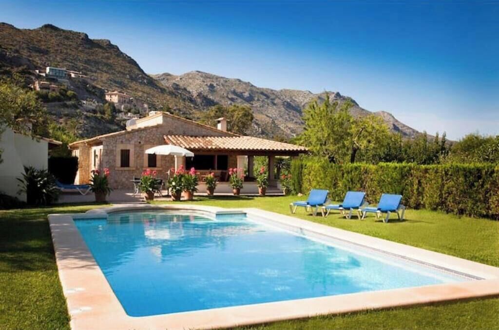 A swimming pool with lounge chairs, a stone house with a tiled roof, and mountains in the background.