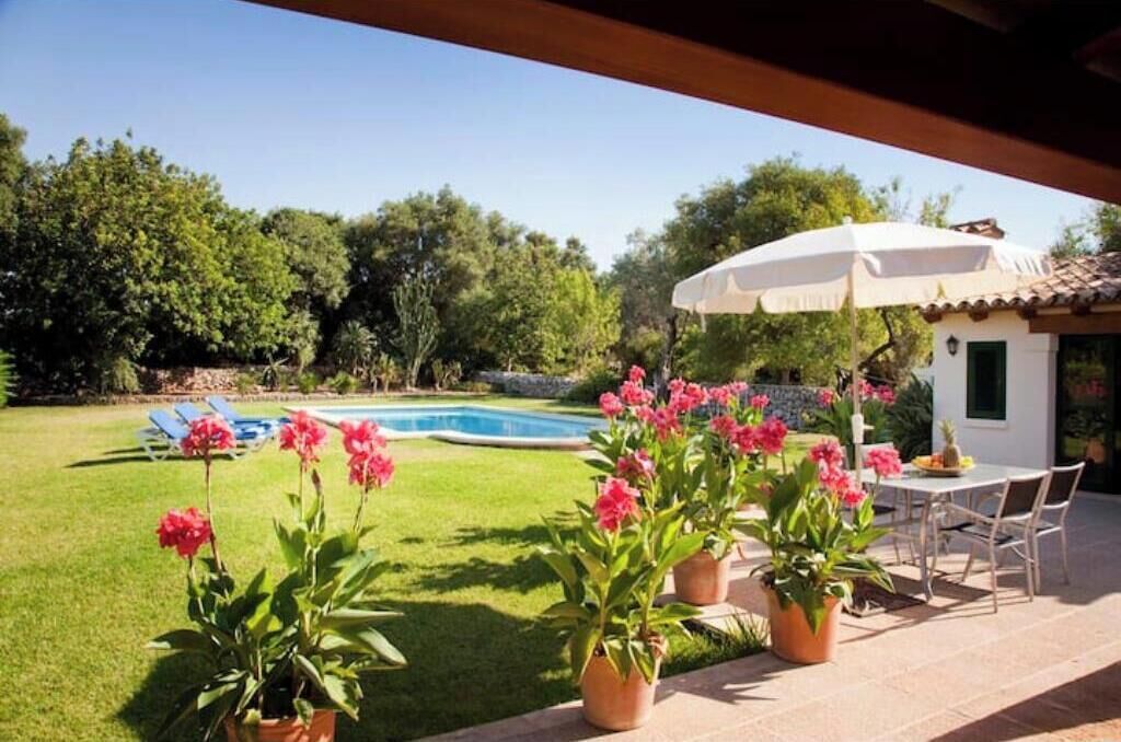 Un área junto a la piscina con una mesa, sillas, plantas en macetas y un paraguas blanco.