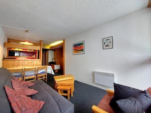 A compact living space with a sofa, a dining table with chairs, a kitchenette, and two framed pictures on the wall.