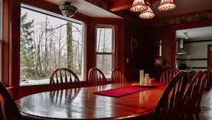 A dining area with a large wooden table and chairs, large windows with a forest view, and a kitchen in the background.